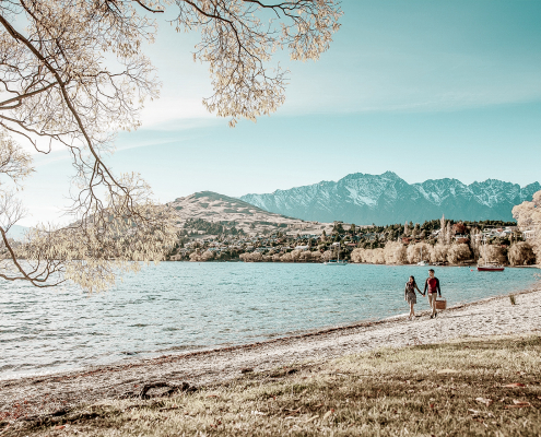 Two people walking on Queenstown Wanaka lakeshore with mountains in background.